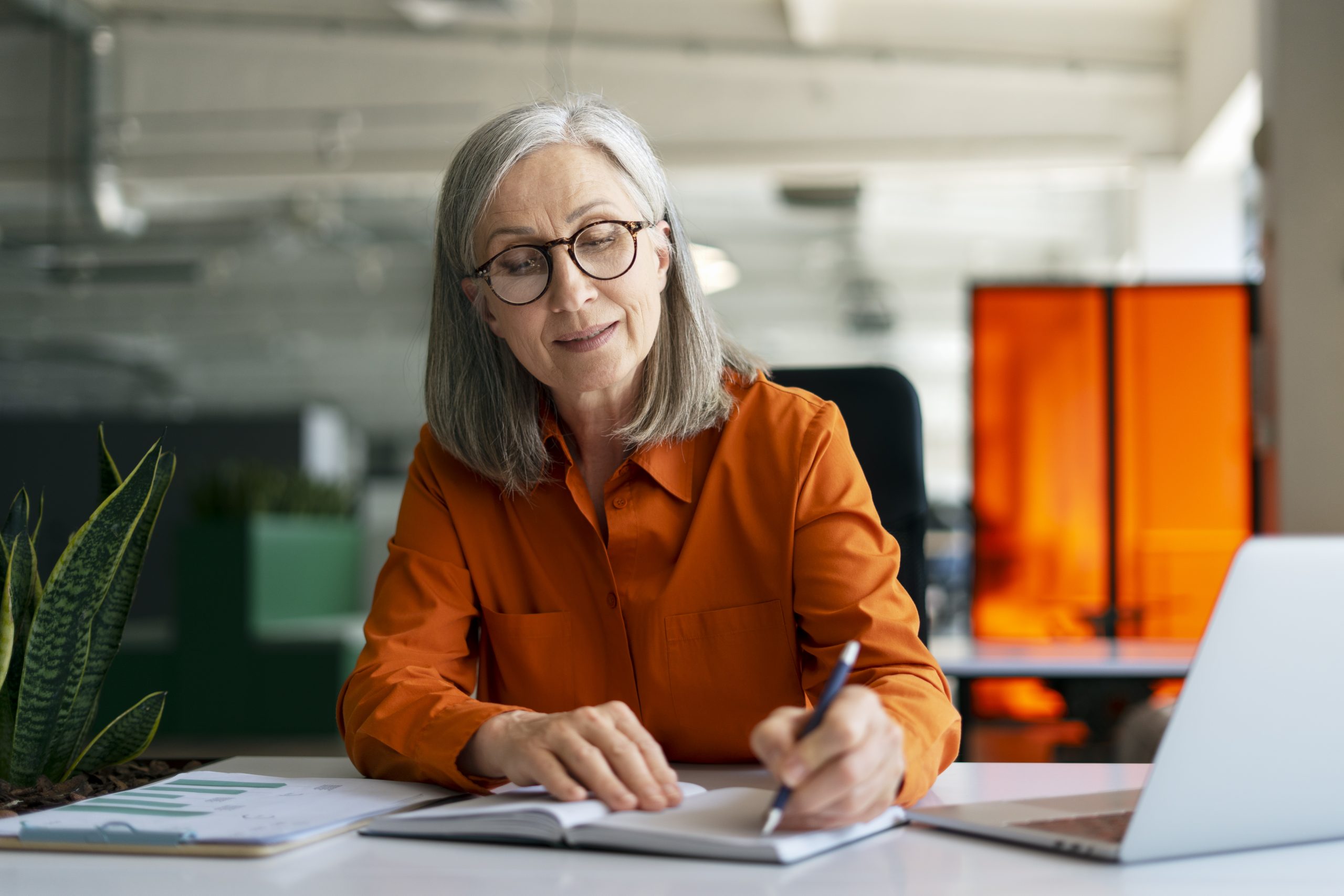 Mature woman, writer using laptop computer, taking notes, working project in modern office. 60 years old female, financier working at workplace. Successful business