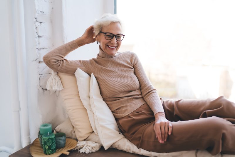 Positive elderly lady with gray hair in casual apparel and eyewear resting on soft blanket and cushions at large window and looking at camera